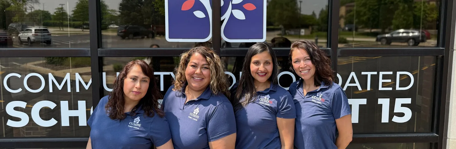 Four smiling women in CCSD15 polo shirts in front of CCSD15 logo'ed window